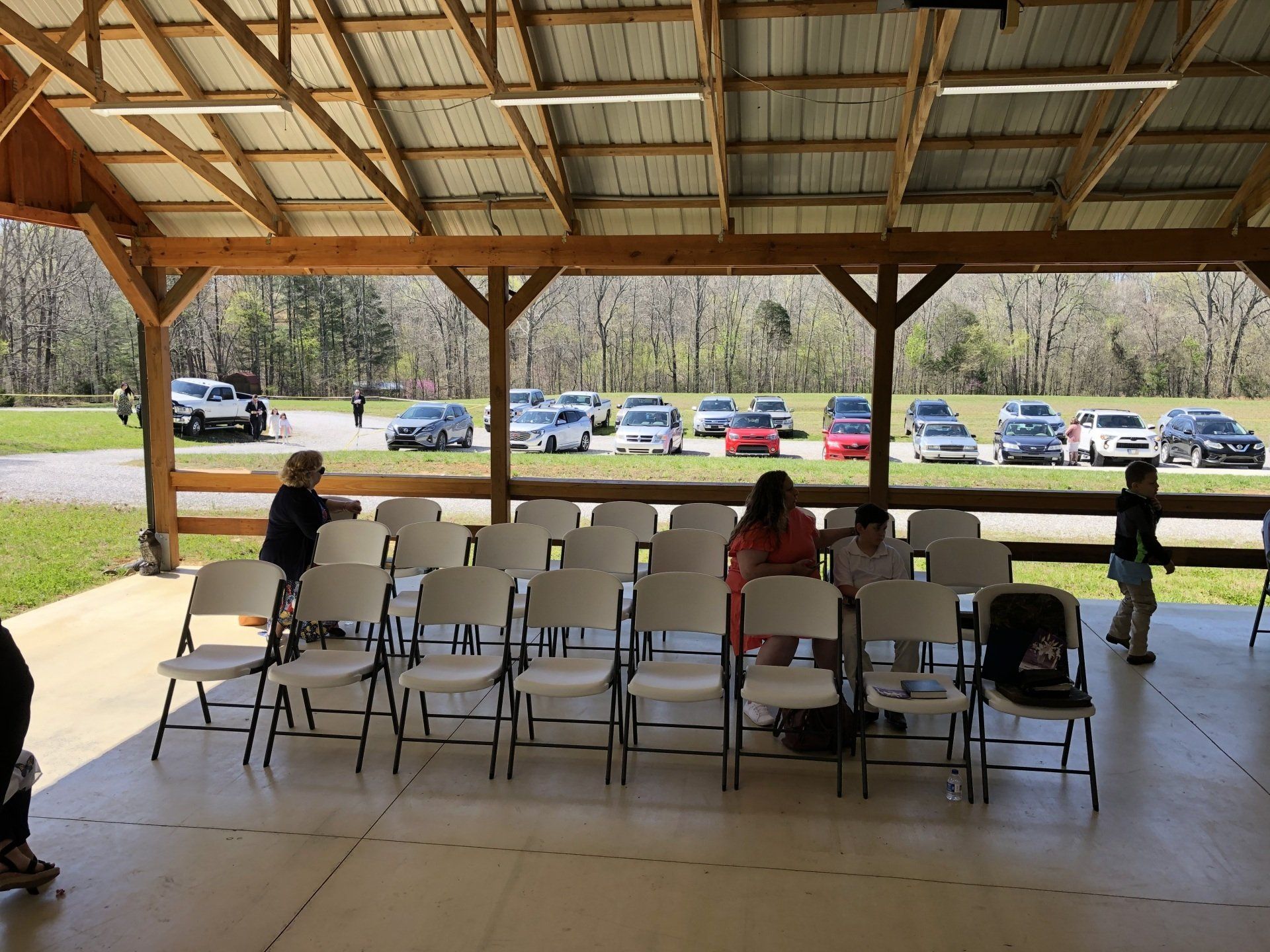 Rows of white chairs set under a pavilion. People mill about. Cars are parked in the background.