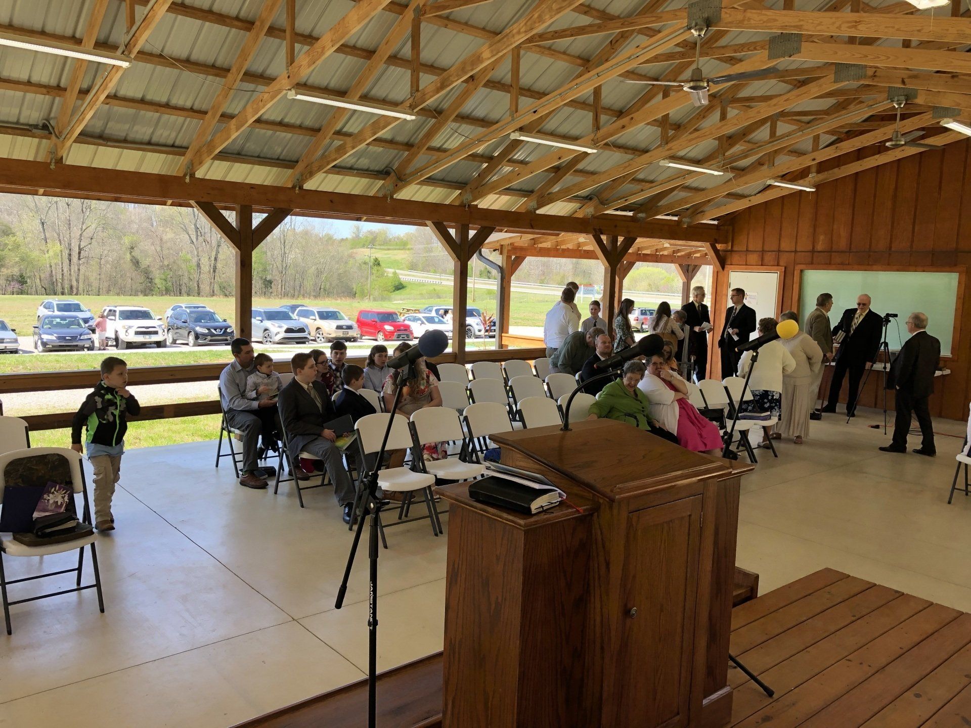 People gathered under a pavilion with a podium and chairs, with cars parked outside.