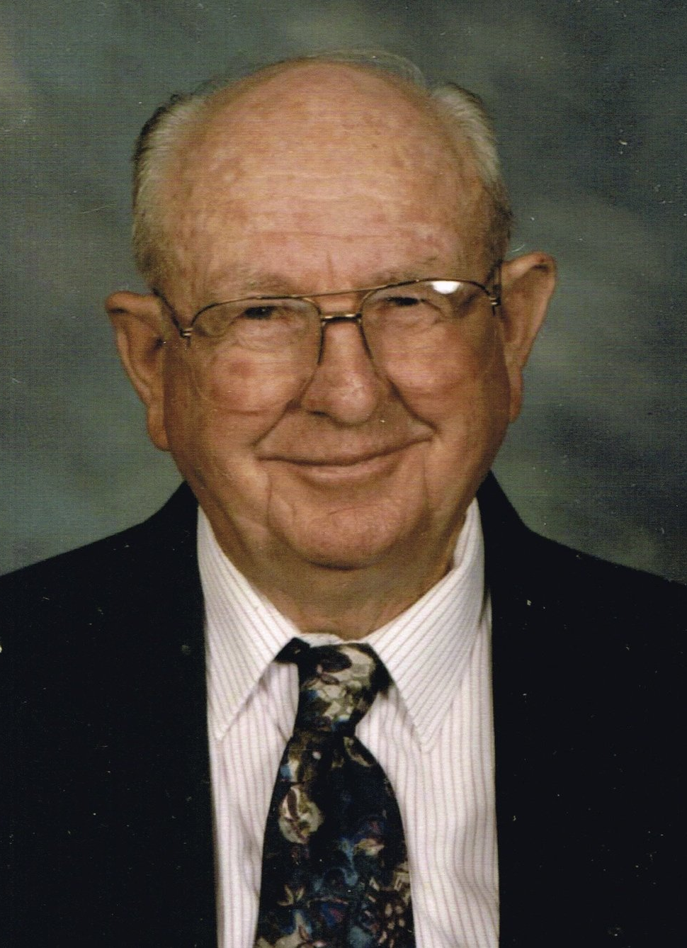 Smiling older man wearing glasses, a suit jacket, and patterned tie.