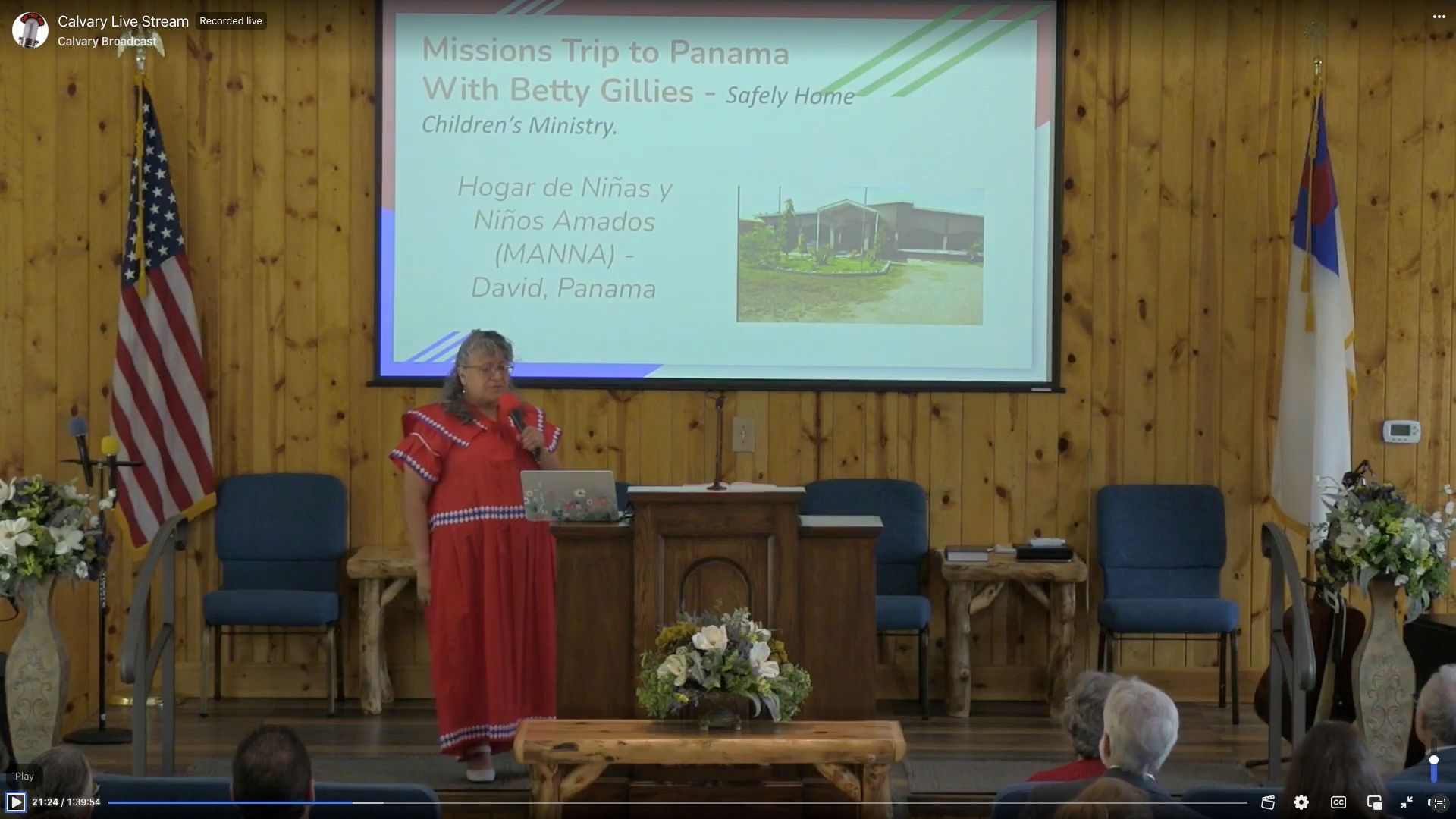Woman presenting in front of a screen displaying a trip to Patmos.  A US flag and church flag are on either side.