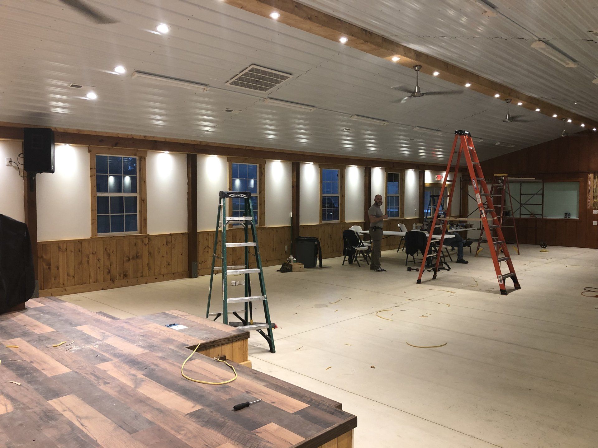 Interior of a large room under construction. Stage in foreground, windows along the side wall, ladders present.