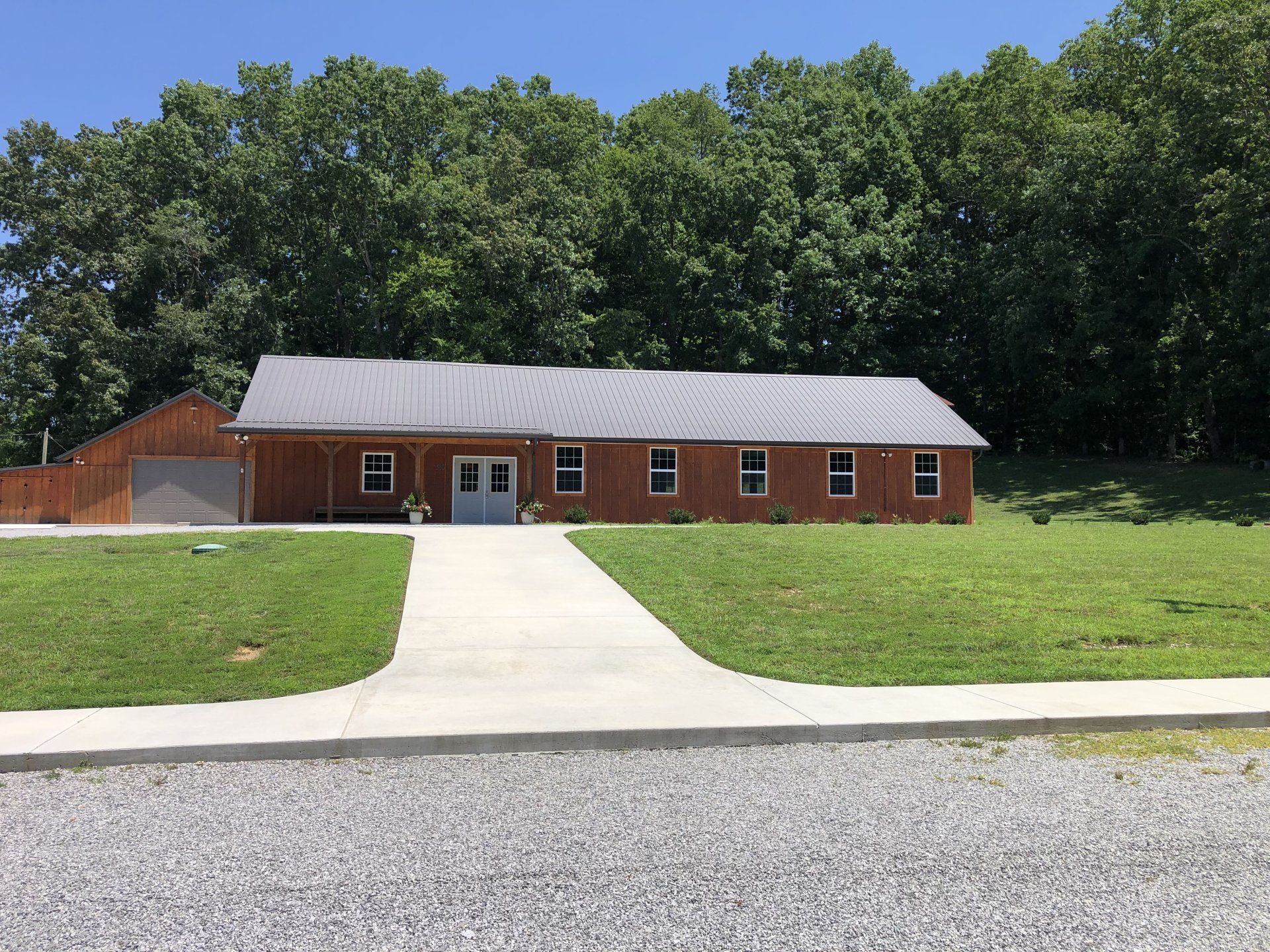 Wooden building with gray roof, windows, and concrete path, in front of a green hill and trees.