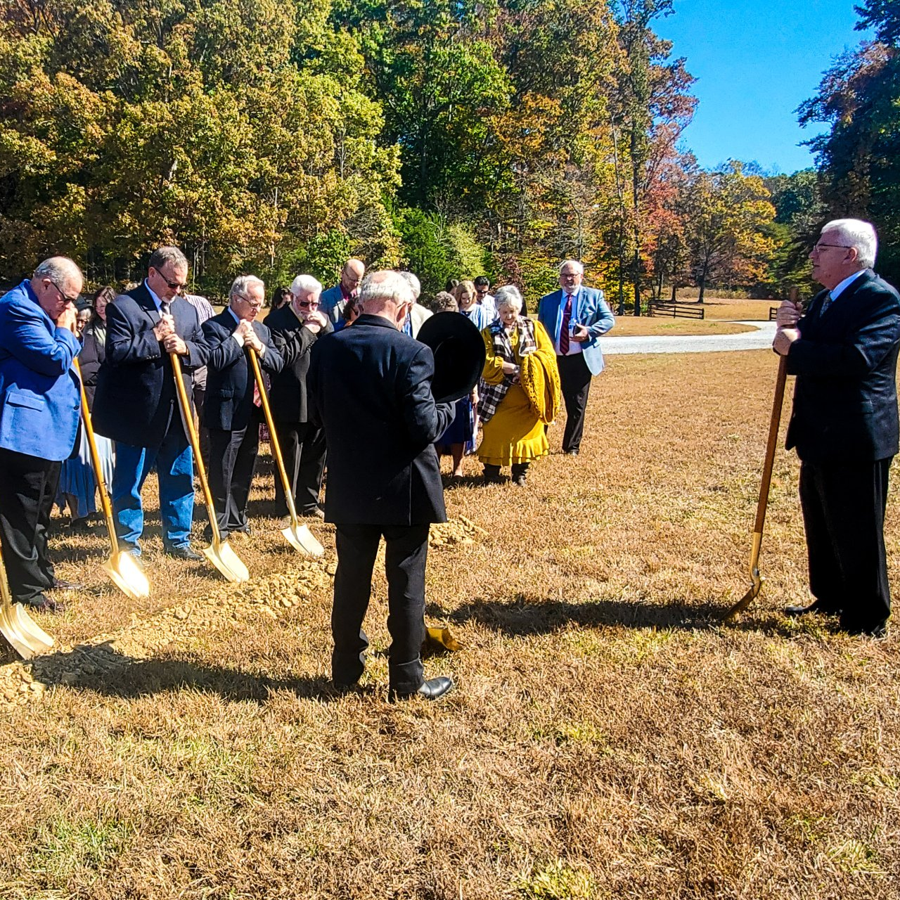Group of people with shovels in a field, listening to a man speaking. Sunny day, trees in background.