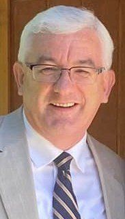Man with glasses, gray hair, light suit, and striped tie smiles broadly outdoors.