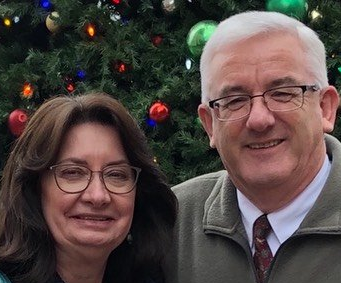 Couple smiling in front of a Christmas tree, both wearing glasses.