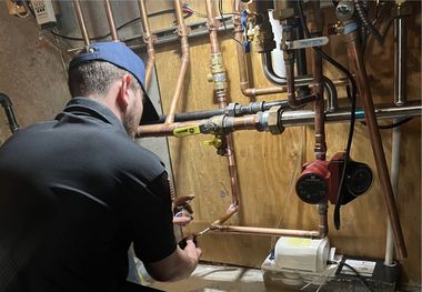 Plumber inspecting copper pipes and valves in a utility room. He wears a blue cap and black shirt.