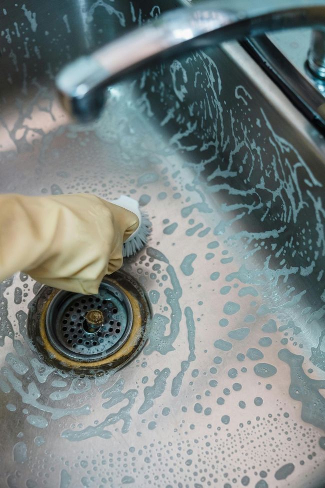 Person wearing glove scrubs a dirty sink drain with a brush, creating suds.