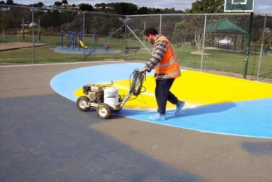 Worker using a specialized machine to apply bright blue and yellow coating on a sport court surface during a court installation service in Chandler, AZ