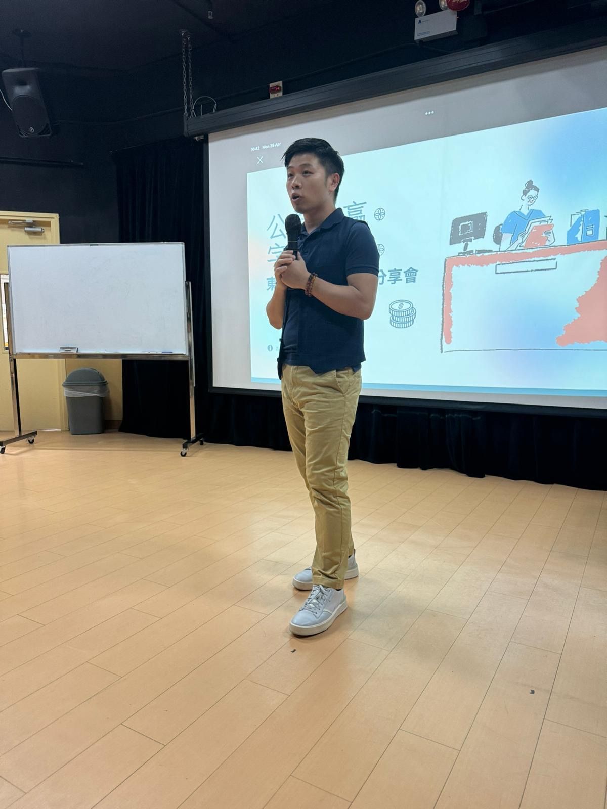 A man in a navy shirt and khaki pants speaks into a microphone in front of a projector screen. He is in a room with a whiteboard and hardwood floors.