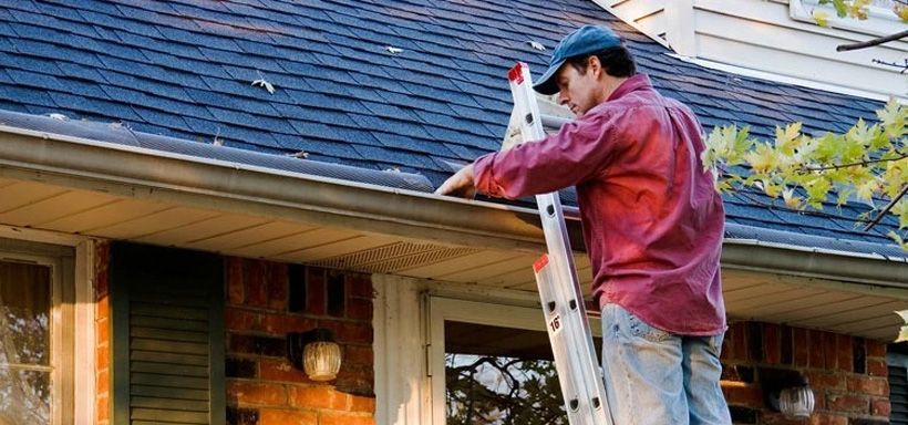 Image of a man in red shirt and jean on a ladder cleaning gutters on home