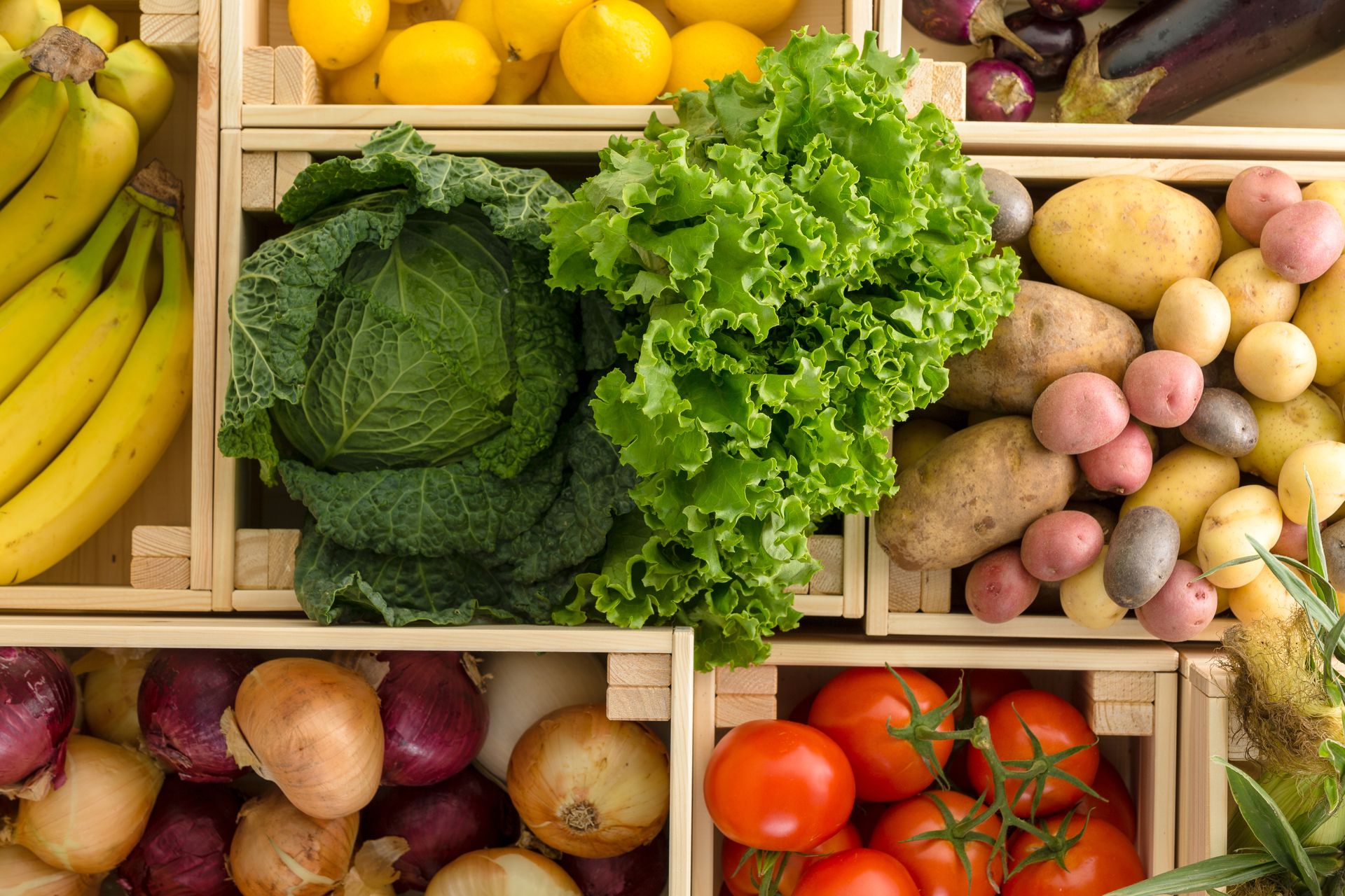 Assorted fresh fruits and vegetables in wooden crates, including bananas, lettuce, potatoes, tomatoes, and onions.