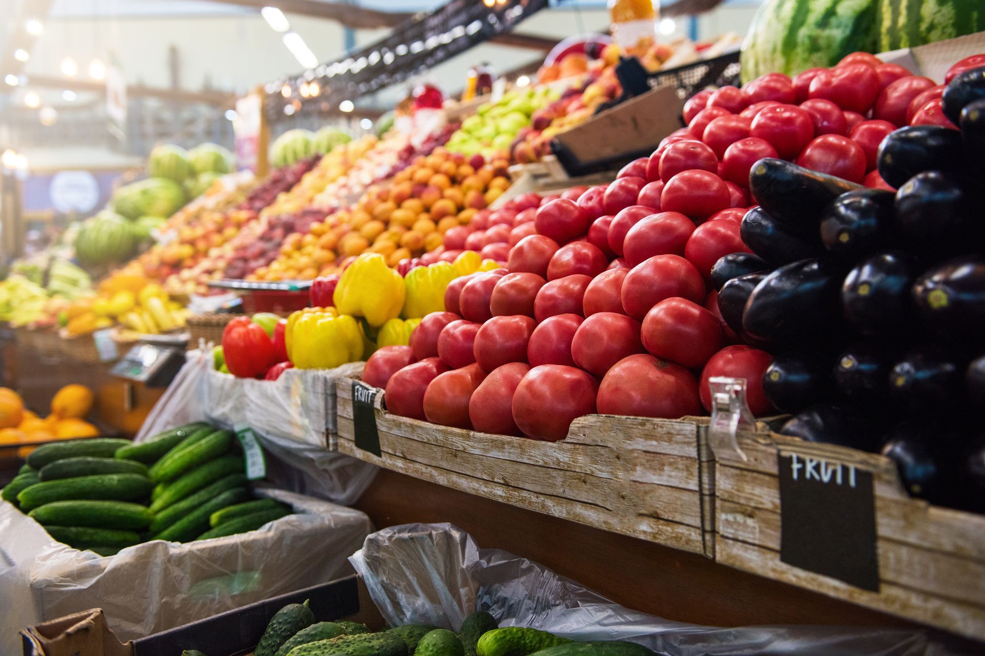 Produce displayed on market stall: red apples, lemons, cucumbers, and glossy eggplants in wooden crates
