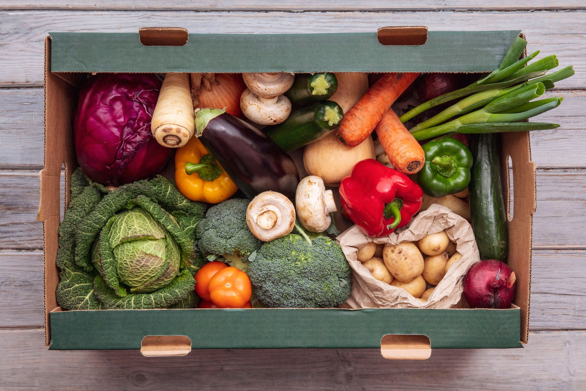 Assorted fresh vegetables in a wooden crate, including peppers, carrots, eggplant, mushrooms, potatoes, and greens