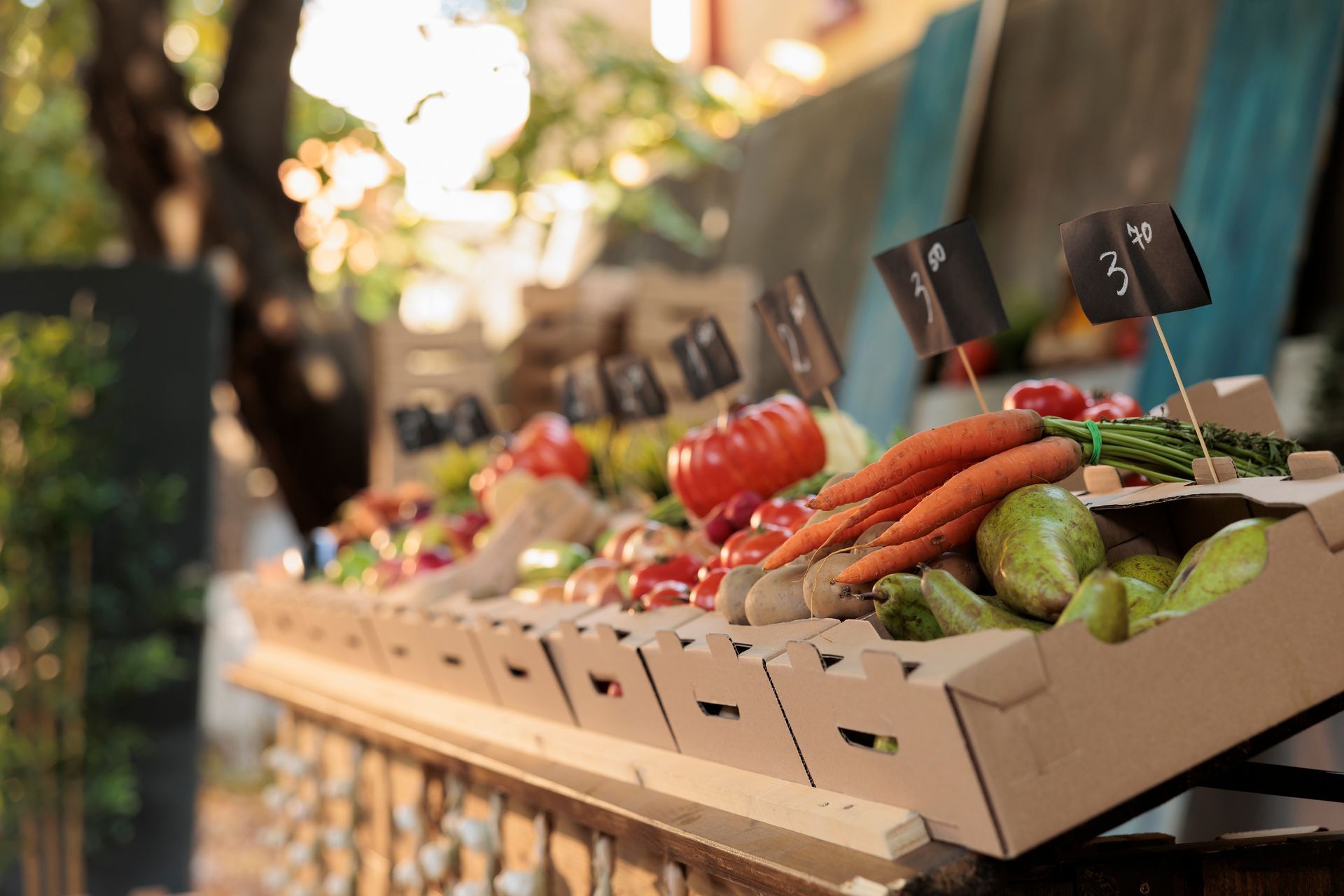 Produce displayed at an outdoor market stall in cardboard boxes, with tomatoes, carrots, and green vegetables on a table.