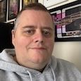 Close-up selfie of a person in a gray hoodie indoors, with framed pictures on the wall behind.