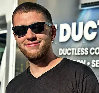 A man wearing sunglasses and a black shirt is standing in front of a ductless company sign.