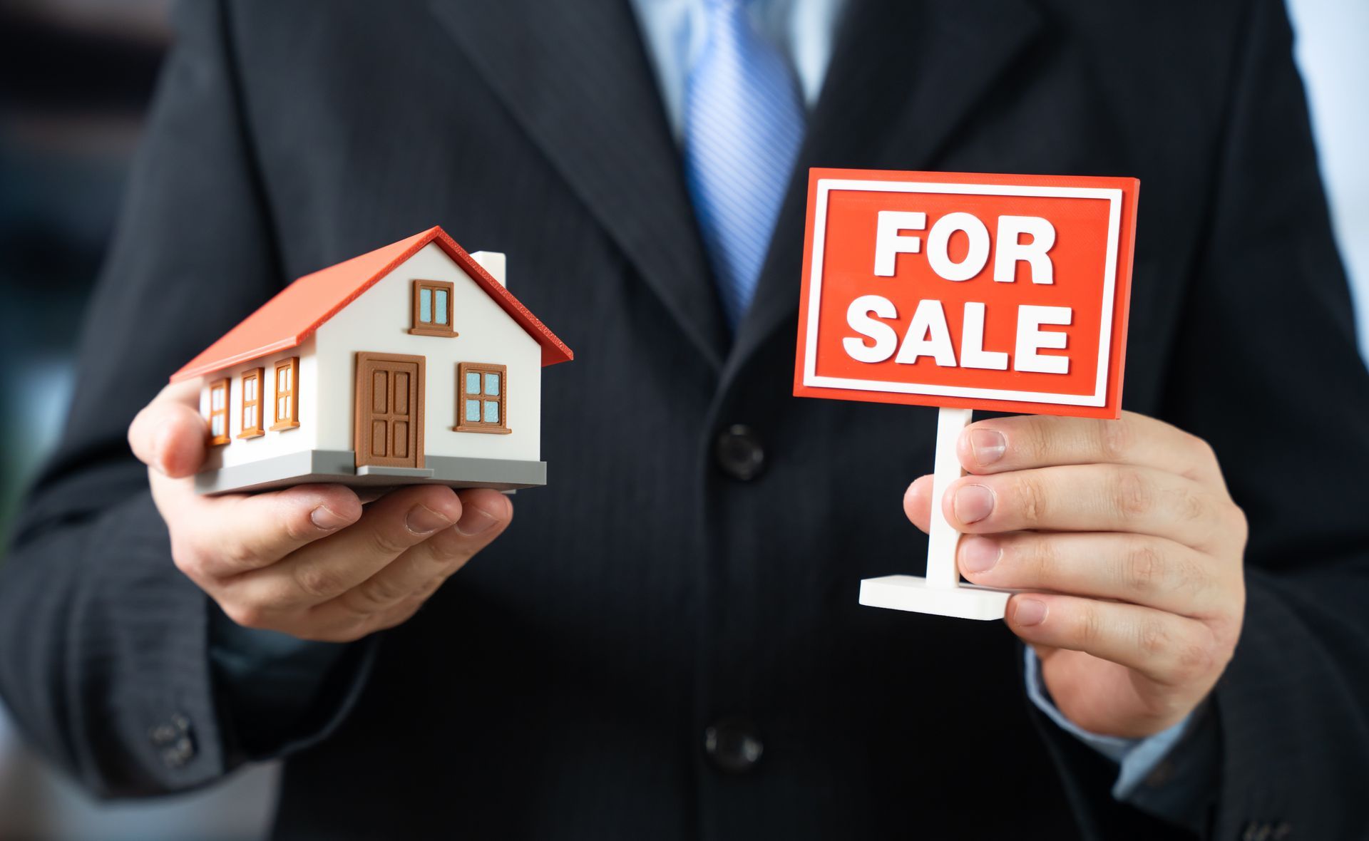 Real estate agent holding a miniature house model and a For Sale sign.