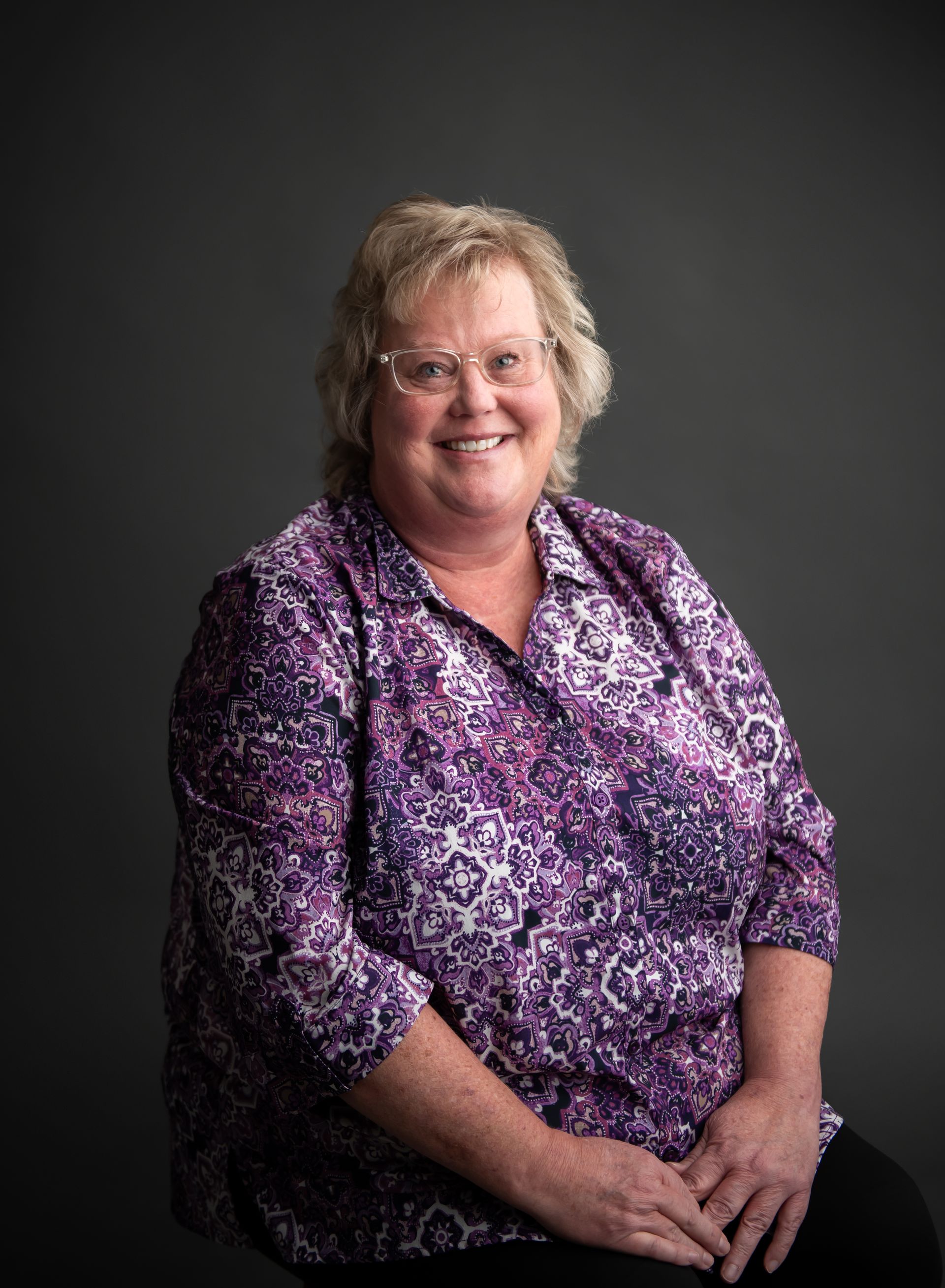 Smiling woman in a purple patterned blouse seated against a dark gray background.