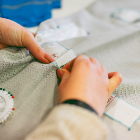 Hands pinning pale fabric pieces together on a sewing table with a measuring tape visible