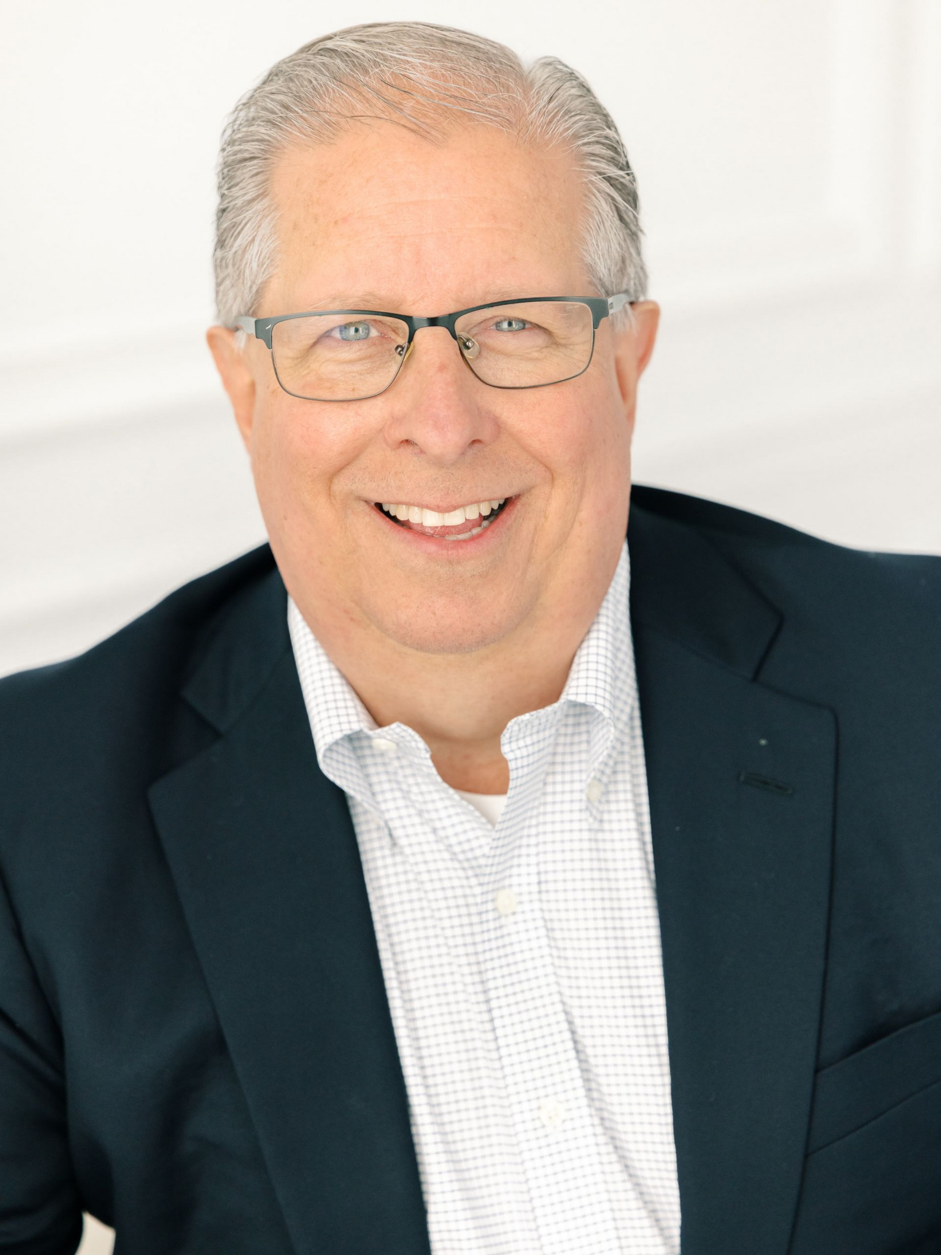 Man smiling, wearing glasses and a dark blazer over a checkered shirt.