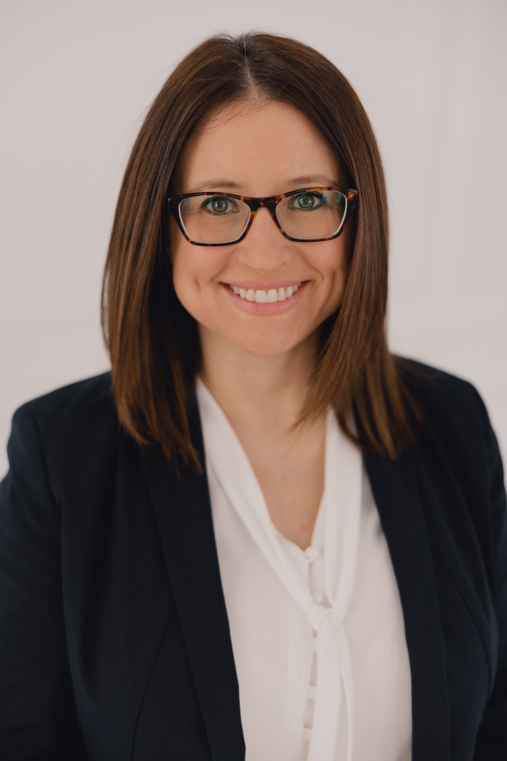 Woman with glasses smiles, wearing a blazer and white blouse, against a white background.
