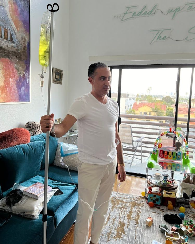 Man in white shirt standing in a bright living room beside a couch and IV stand