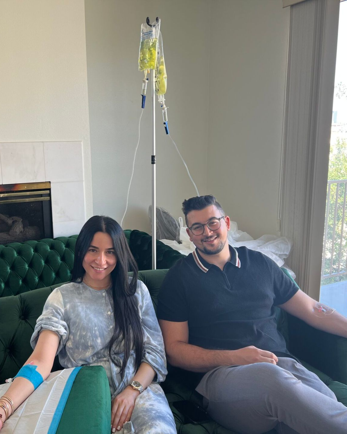 Two people sit on a teal couch in a bright room, smiling near an IV stand.