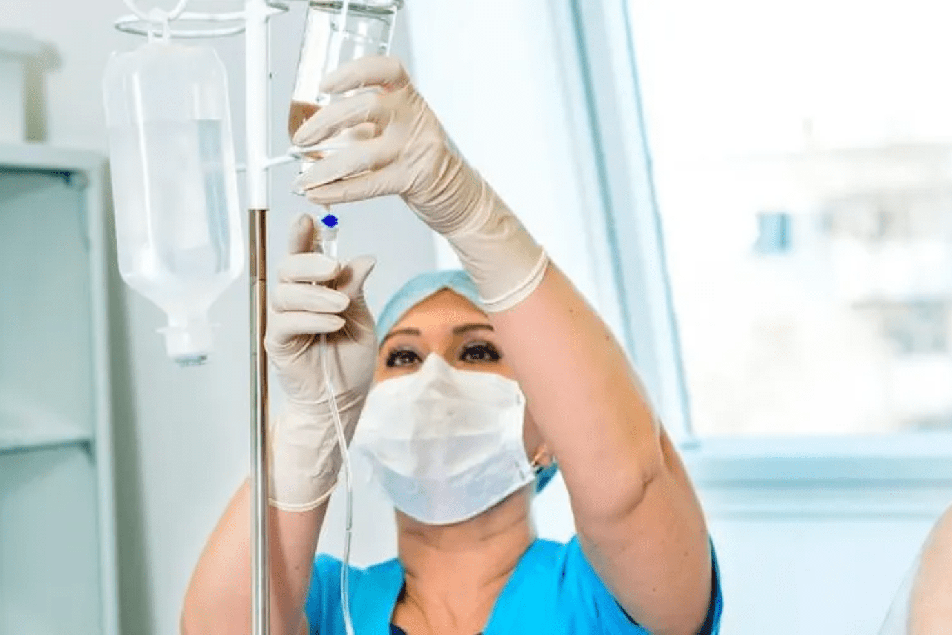 Nurse adjusting IV drip in a hospital room, wearing mask, cap, and gloves.