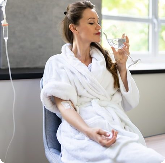 Woman in a white robe sitting in a chair, holding a glass of water with an IV line in her arm.