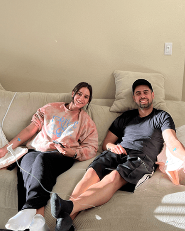 Two people relaxing on a beige couch in a sunlit room, smiling and sitting casually with pillows.