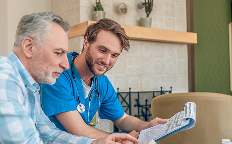 A healthcare worker in blue scrubs smiles as he reviews paperwork with an older patient indoors.