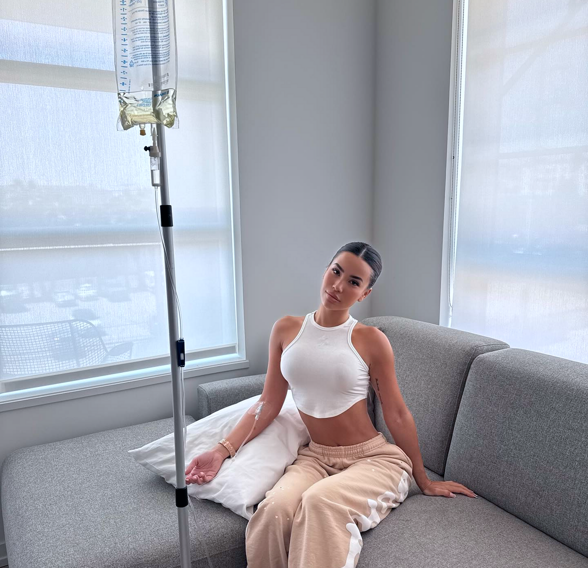 Woman on a couch, receiving IV fluids; wearing white top and tan pants, light room.