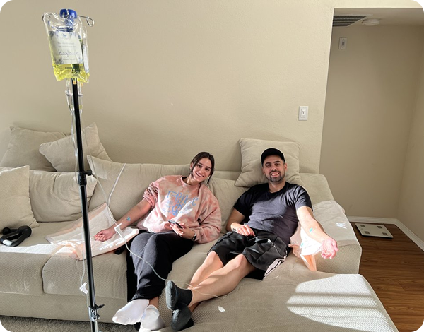 Two people reclining on a beige couch with IV bags, in a sunlit living room.