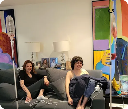Two people sit on a couch in a colorful art-filled room, smiling beside lamps and framed photos.