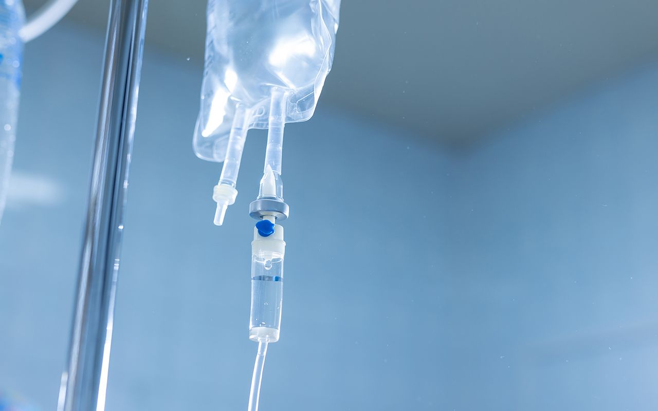 An IV fluid bag and drip chamber hanging on a stand against a blue clinical background.