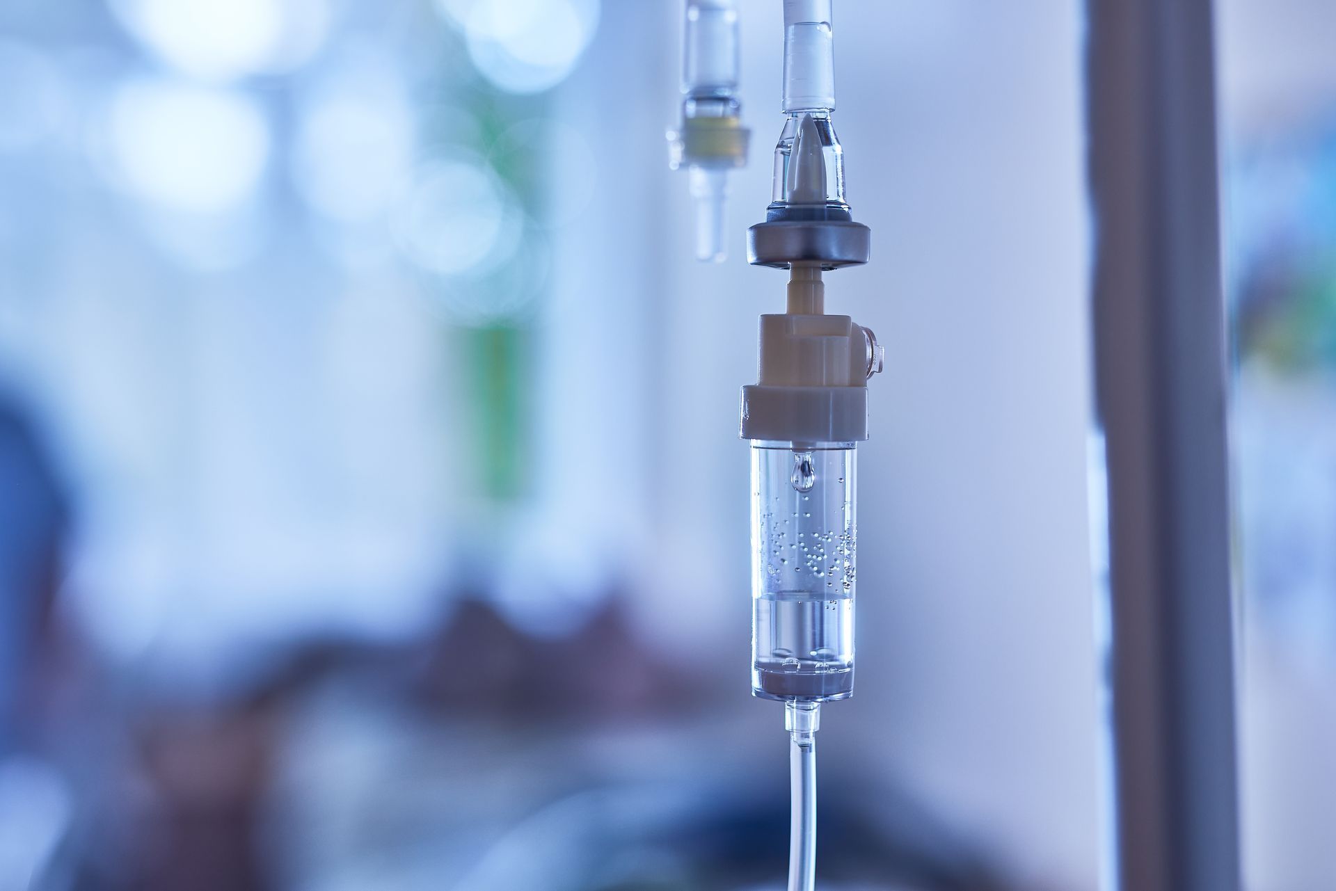 Close-up of a medical intravenous (IV) drip chamber with clear fluid, set against a softly blurred hospital background.