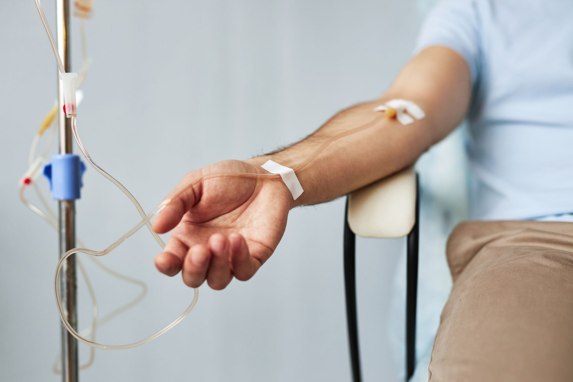A person's arm resting on a chair, connected to an intravenous (IV) drip stand with tubing and medical tape.