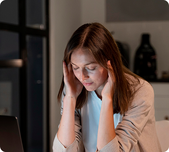Woman with hands on head, appearing stressed, at a laptop in a dimly lit room.