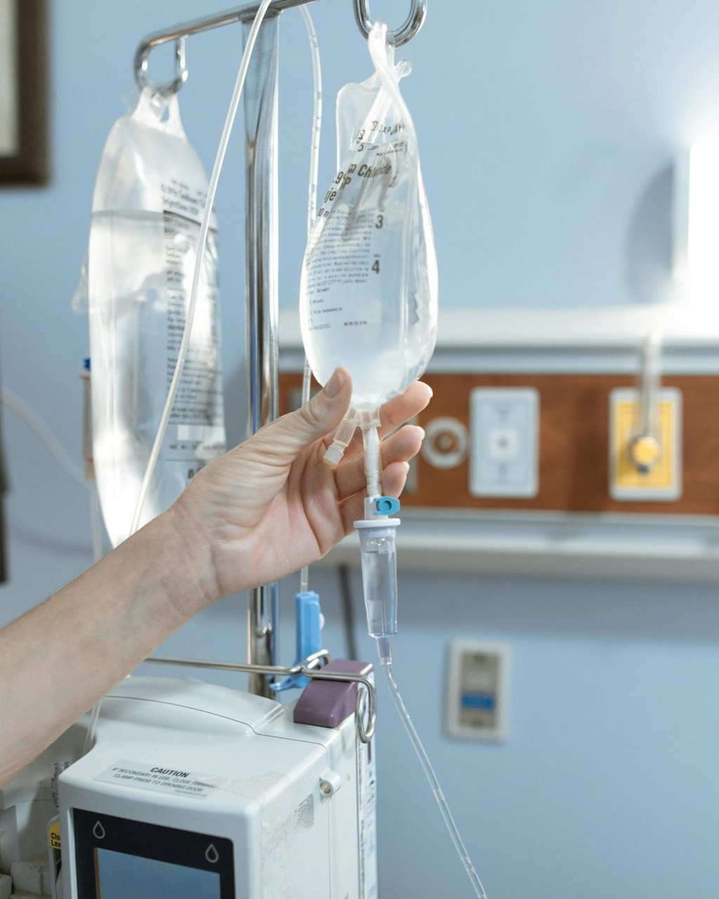 Hospital IV bags hanging beside a patient monitor, with a hand adjusting the infusion line.