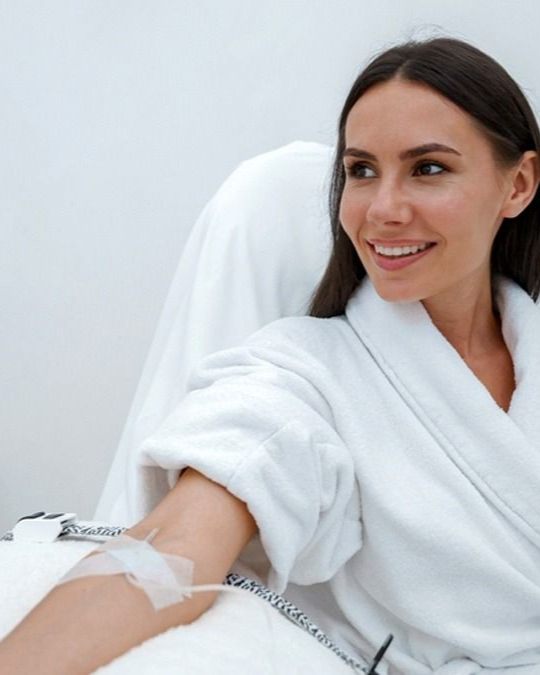 Woman in white robe smiling during an arm treatment, with a small bandage on her forearm
