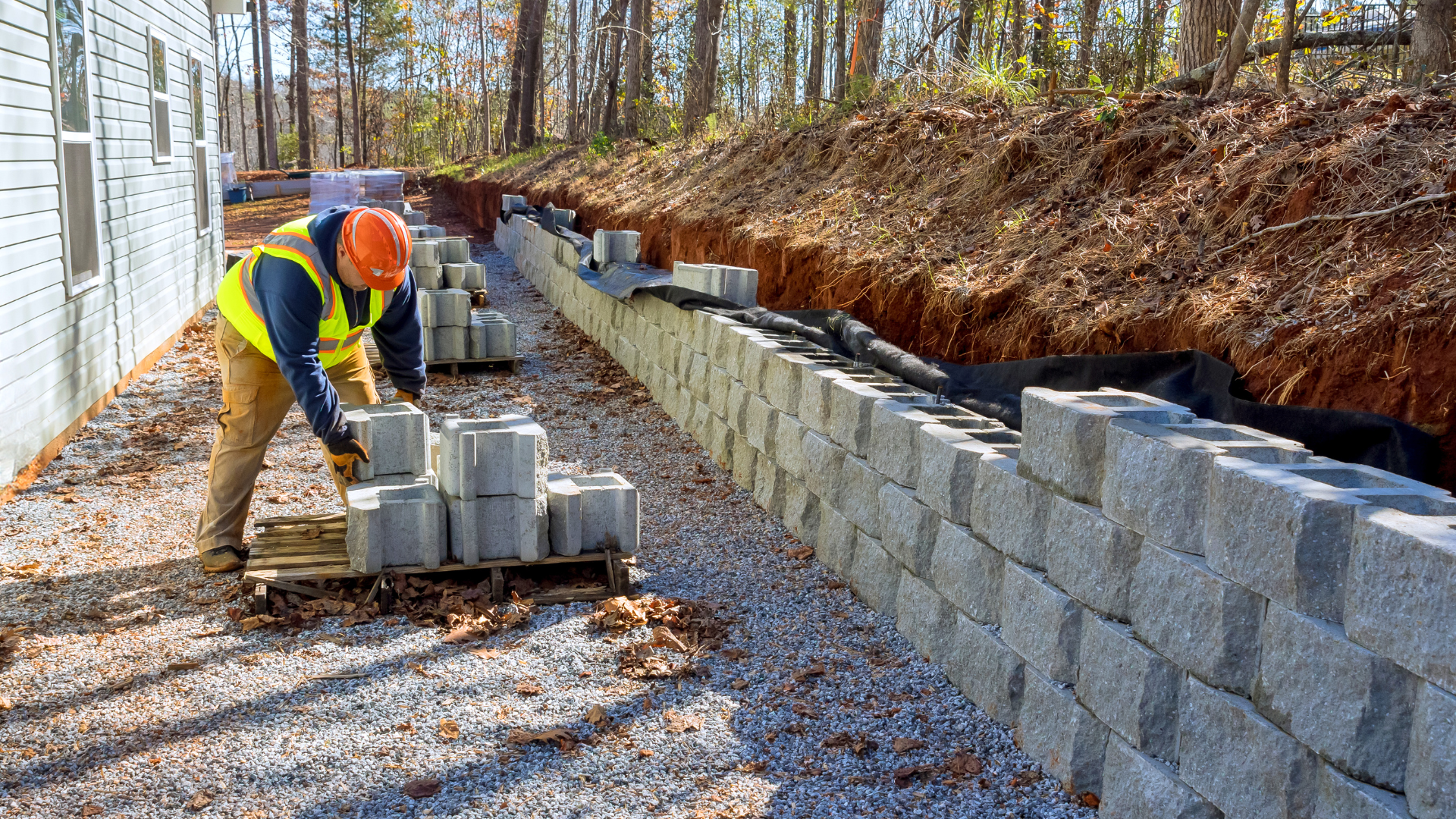 Worker stacking cinder blocks beside a retaining wall at a wooded construction site