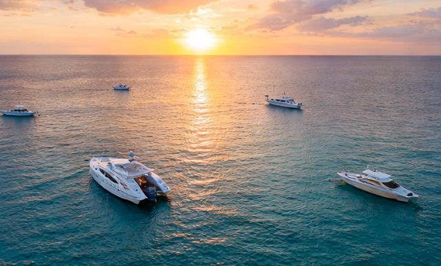Several white yachts float on calm, sunlit ocean waters during a golden sunset.