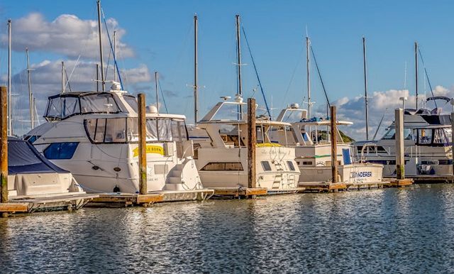Several white boats are docked in a marina under a bright blue sky with scattered clouds.