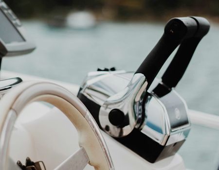 Chrome and black boat throttle controls mounted on a white console, with a steering wheel and water in the background.