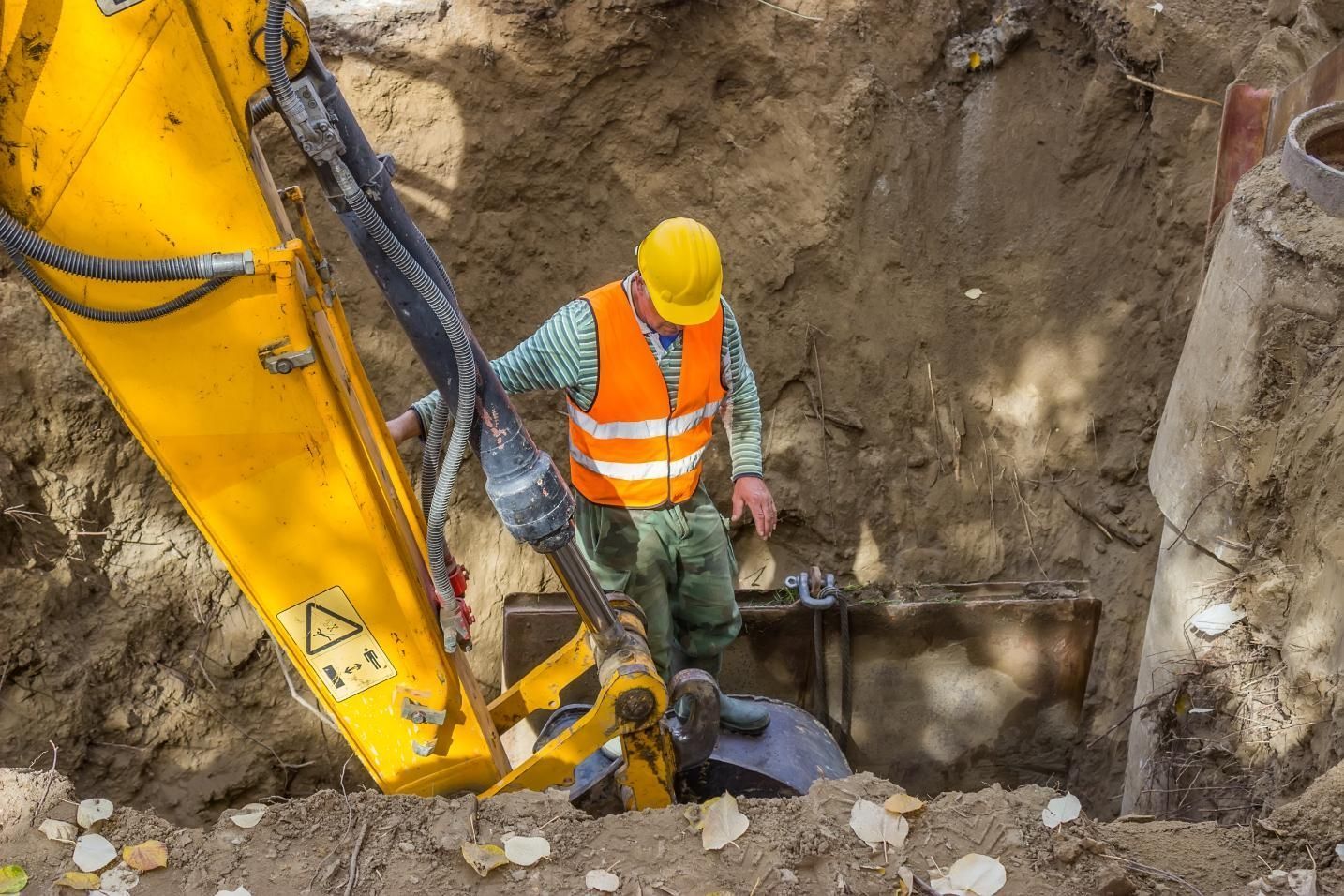 Construction worker in orange vest and yellow hard hat standing beside excavator in a dirt trench