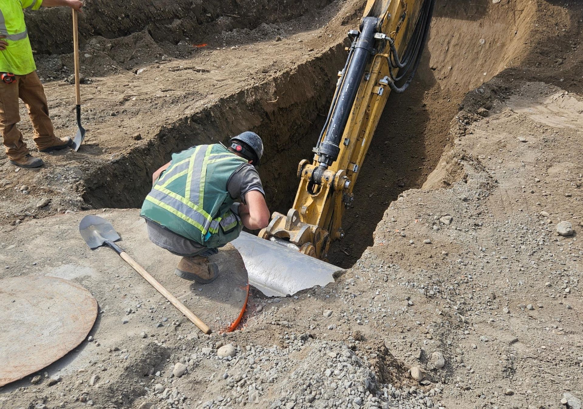 Worker in reflective vest crouches by an excavator in a dirt trench, guiding a metal sheet.