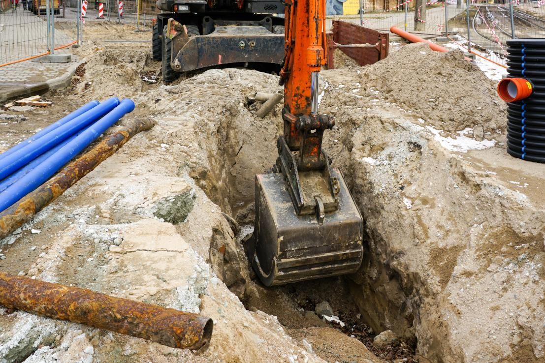 Excavator digging a trench at a construction site beside blue pipes and dirt piles