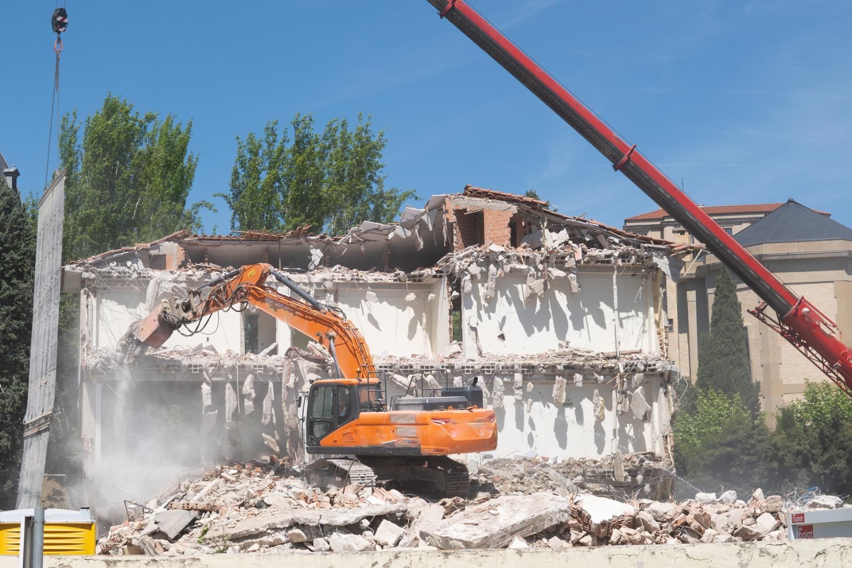 Orange excavator demolishing a partially collapsed building, with debris and dust under a clear blue sky