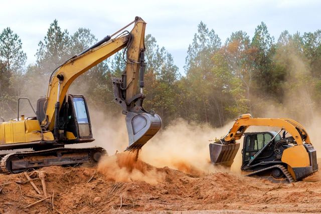 Two excavators digging red dirt, kicking up dust in a wooded work site.