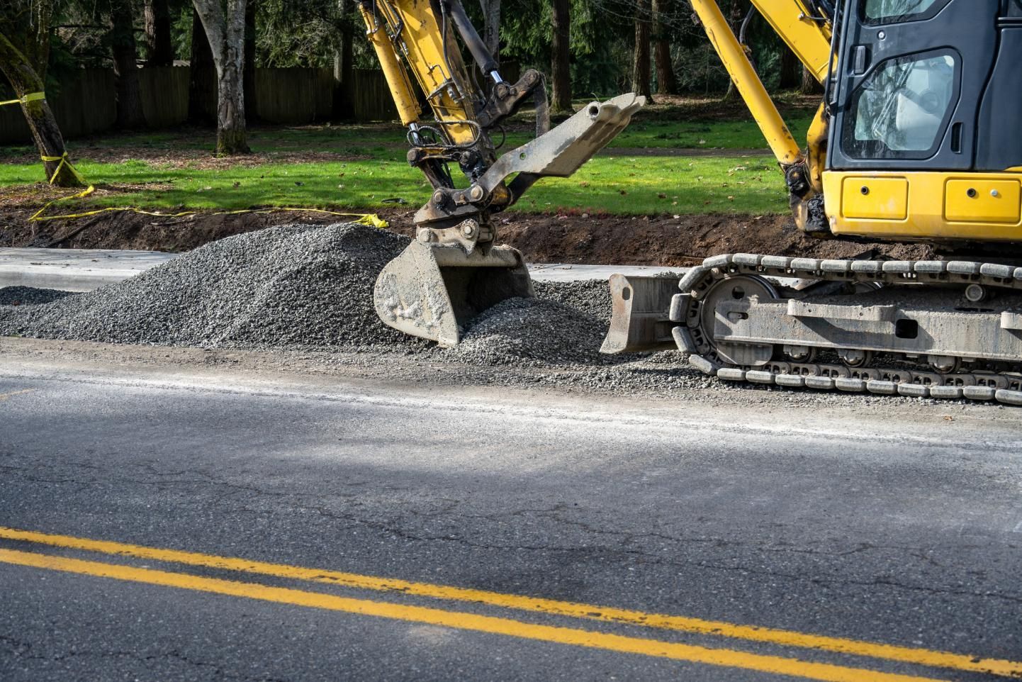 Yellow excavator spreading gravel beside a road with double yellow lines.